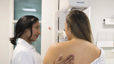 Image of a women getting a mammogram, supported by a nurse. Image is showing the back of the patient with a particular focus on the machine. 